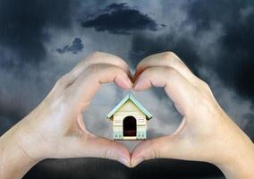 manos de la persona haciendo una forma de corazón con una casa de madera en el fondo de las nubes de lluvia foto