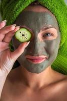 Beautiful young woman with facial mask on her face holding slices of cucumber. photo