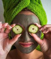 Beautiful young woman with facial mask on her face holding slices of cucumber. photo