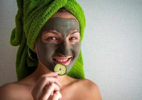 Beautiful young woman with facial mask on her face holding slices of cucumber. photo