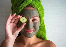 Beautiful young woman with facial mask on her face holding slices of cucumber. photo