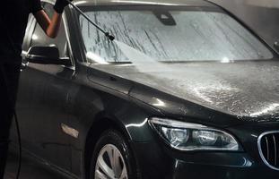 utilizando equipos con agua a alta presión. el automóvil negro moderno es limpiado por una mujer dentro de la estación de lavado de autos foto