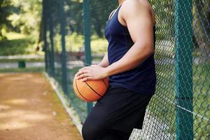 African american man takes a break and leaning on the metal mesh with ball on the court outdoors photo