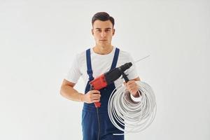 con taladro y cables en las manos. trabajador de sexo masculino en uniforme azul de pie dentro del estudio contra el fondo blanco foto