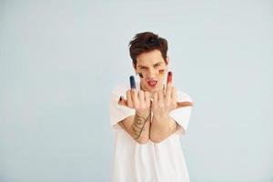 Young gay man is standing in the studio and posing for a camera. Shows middle fingers photo