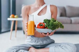 With healthy food and orange juice in hands. Young woman with slim body shape in sportswear have fitness day indoors at home photo