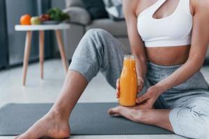 Holds glass with orange juice in hands. Young woman with slim body shape in sportswear have fitness day indoors at home
