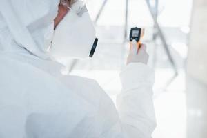 Female doctor scientist in lab coat, defensive eyewear and mask standing indoors with infrated thermometer photo