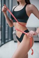 Slender woman in black fitness underwear standing indoors in room at daytime with measuring tape photo