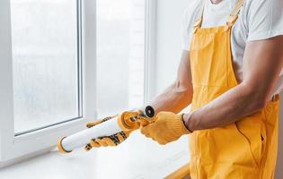 Handyman in yellow uniform works with glue for window indoors. House renovation conception photo