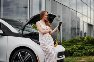 Looks at device. Woman on the electric cars charge station at daytime. Brand new vehicle photo