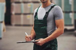 Storage worker in green colored uniform and notepad in hands checks production photo