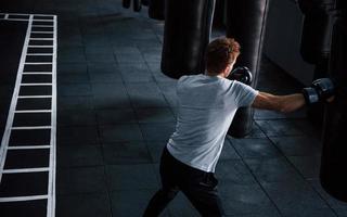 Young man in white shirt and boxing protective gloves doing exercises in gym with pushing bag photo