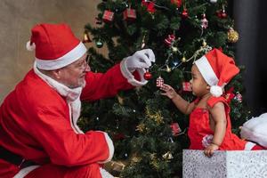 African American baby is getting ornamental bauble as present from Santa claus at night by the fully decorated christmas tree for season celebration photo