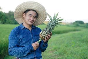 Handsome Asian man farmer wears hat, blue shirt, holds pineapple fruit. Concept , Agriculture crop in Thailand. Farmer satisfied. Organic crops. Seasonal fruits. photo