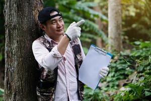 Asian man botanist is at forest, holds paper clipboard, point to something to analysis and research about  trees. Concept, forest valuation. Conservation of environment. photo