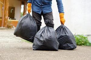 Closeup man holds black plastic bag that contains garbage inside, stand in front of house. Concept , Waste management. Environment problems. Daily chores. Throw away rubbish . photo