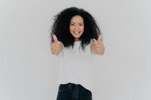 Happy young woman with Afro hairstyle, shows thumbs up, accepts or agrees awesome plan, gives her approval, smiles broadly, stands indoor, says excellent decision, isolated over white background photo