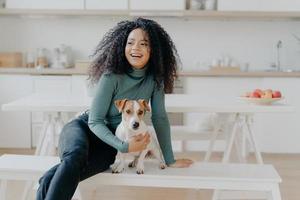 Joyful Afro woman sits at white bench together with dog against kitchen interior, table with plate full of red apples, get pleasure while playing at home. Animal owner feels care and responsibility photo