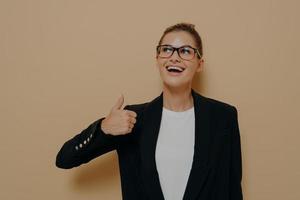 Portrait of satisfied female in eyeglasses smiling while showing thumb up with hand photo