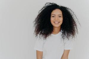 Headshot of smiling woman has bushy luminous hair, stands confident and pleased, minimal makeup, enjoys lively small talk, dressed in comfortable white t shirt, poses indoor. Happy emotions. photo