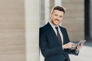 Experienced male economist in formal clothes works on digital tablet, has work break, stands in office interior with copy space. Successful entrepreneur prepares business report on modern device photo
