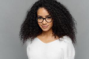 Close up shot of dark skinned female has satisfied expression, bushy curly hair, wears spectacles and white jumper, looks straightly at camera, models over grey background. Natural beauty concept photo