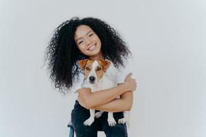 Horizontal shot of pleasant looking curly young woman embraces lovely pet, holds cute dog, smiles broadly, tilts head, isolated against white background. People, animals, good emotions concept photo