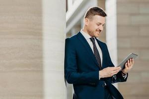 El exitoso trabajador de oficina masculino con traje negro usa una tableta moderna para el trabajo a distancia, conectado a Internet inalámbrico, posa en un interior moderno, concentrado en la pantalla. concepto de tecnología foto