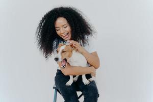 una alegre mujer afroamericana juega con un perro pedigrí, vestida con ropa informal, acariciando a su mascota favorita, aislada sobre fondo blanco, sentada en una silla. expresiones humanas positivas. concepto de amistad foto