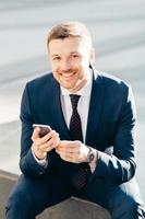 Positive successful male CEO sits outdoor, uses modern smart phone for reading news in message, checks notification, dressed in elegant suit, has satisfied expression, enjoys online communication photo