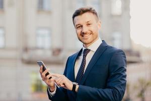 Cheerful male financier holds modern cell phone chats with partners, arrange business meeting, poses outdoors, has happy expression, dressed in black elegant suit. Successful businessman reads message photo