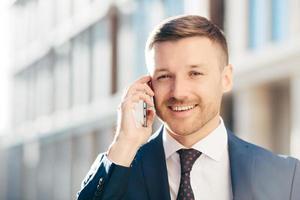 Outdoor shot of prosperous successful unshaven businessman wears formal clothes, has pleasant talk with business partner via cell phone, discuss future meeting, poses against blurred background photo