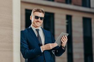Portrait of happy male entrepreneur sends text message to colleague via touch pad, connected to wireless internet, updates software, wears trendy sunglasses and black suit. People and technology photo