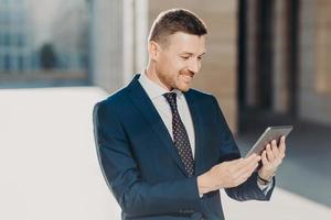 Cheerful male manager reads notification on digital tablet, wears formal black suit with tie, connected to wireless internet, watches video on web page. Business and modern technologies concept photo