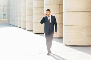 Photo of happy satisfied businessman makes telephone conversation, lools down, strolls near office building, uses roaming, dressed in formal clothes. People, business and communication concept