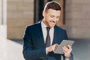 Confident handsome male manager checks email on tablet computer, updates database on touch pad, wears elegant formal clothing, connected to wireless internet, poses outdoor. Business concept photo