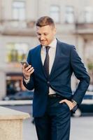 Vertical shot of prosperous male manager wears elegant suit, keeps hand in pocket, uses smart phone, being always in touch, reads business news on internet website, concentrated into screen. photo