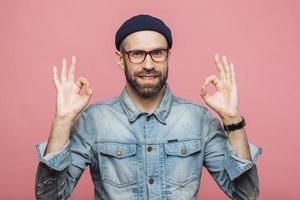 Cheerful bearded male in fashionable clothing, makes ok sign, being satisfied with something, isolated over pink studio background. Unshaven middle aged man gestures indoor, shows his approval photo