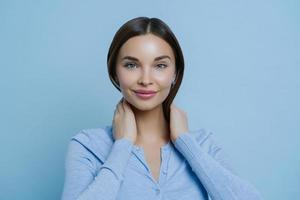 una foto en la cabeza de una joven mujer encantadora mantiene las manos en el cuello, tiene apariencia europea, feliz de escuchar palabras agradables, usa un suéter informal, aislada sobre un fondo azul. concepto de expresiones faciales humanas