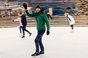 Happy male waves with hand as notices his friend on skating ring, have desire to skate together, rejoices meeting, has good time and mood, entertains himself. People and winter activities photo