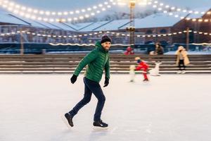 Professional male skater shows his skating talents, being sure on ice ring, looks delightfully in camera, demonstrates his nice skills. Smiling happy man on skating ring. Leisure and professionalism photo