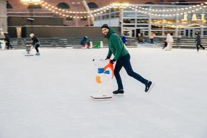 Cheerful man spends free time on skate ring, uses skate aid, learns how to skate, has pleased happy expression, surrounded with people of different age. People, winter, season, hobby concept photo