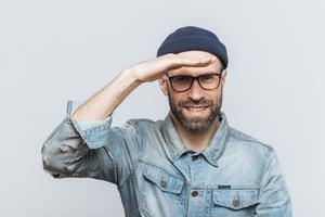 Cheerful unshaven middle aged male with thick stubble looks happily into distance, keeps hand near orehead, tries to see something, wears eyewear and denim jacket, isolated over grey background. photo