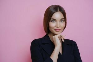 Photo of attractive well groomed businesswoman wears elegant black costume, keeps hand under chin, looks calmly at camera, has gentle look, going to have business meeting isolated over rosy background