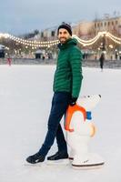 Portrait of cheerful bearded male stands near skate aid, going to skate for first time, being in good mood, enjoys winter holidays. Happy unshaven man in warm clothes and skates stand on ice rink photo