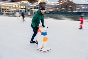 Outdoor shot of unexperienced male skater uses skate aid for go skating on ice, being in good mood, enjoys spending free time with friends during wonderful winter weather. Leisure concept photo