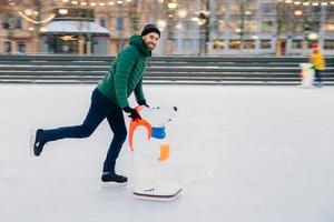 Outdoor shot of happy delightful man with thick beard and mustache looks gladfully into camera, demonstrates going skating on ice ring with skate aid, glad to go in for sport in such way photo