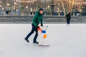 Positive smiling attractive man in warm winter clothes and skates, practices go skating on ice, uses skate rink, looks delightfully into camera, spends Sunday outdoor during winter period photo