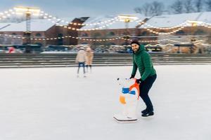 Shot of glad bearded male spends time outdoor on fresh air, enjoys frosty winter weather, goes skating on ice ring, uses skae aid as afraid to fall down. Handsome man has fun on skating ring photo